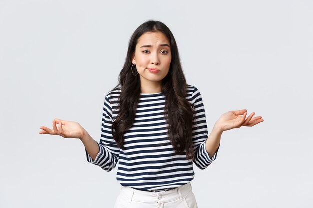 Estilo de vida, belleza y moda, concepto de emociones de personas. Sonriente linda chica despistada no tiene idea, no responde, encogiéndose de hombros con las manos extendidas hacia los lados, de pie fondo blanco