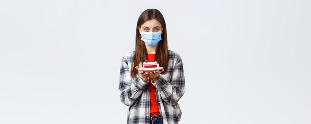 Estilo de vida del brote de coronavirus durante el distanciamiento social y el concepto de celebración de vacaciones Niña linda con máscara médica y atuendo informal sosteniendo pastel de cumpleaños y mirando una cámara seria