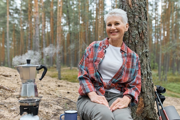 Estilo de vida de camping en el bosque. Alegre mujer europea de mediana edad sentada en el suelo debajo de un pino que va a hacer té, agua hirviendo en una tetera en el quemador de la estufa de gas, con una expresión facial alegre y feliz