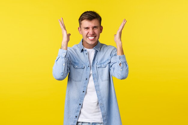 Estilo de vida, emociones de la gente y concepto de ocio de verano. Hombre guapo rubio molesto y cabreado volviéndose loco, perdiendo los estribos, levantando las manos y haciendo muecas de irritación, fondo amarillo.