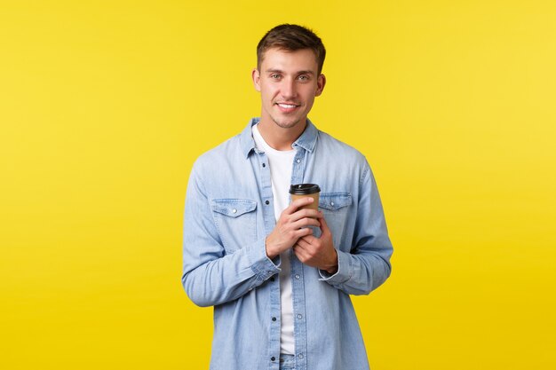 Estilo de vida, emociones de las personas y concepto de ocio. Apuesto joven con sonrisa blanca, pedir café para llevar en la cafetería, beber de un vaso de papel, tener una conversación informal sobre fondo amarillo.