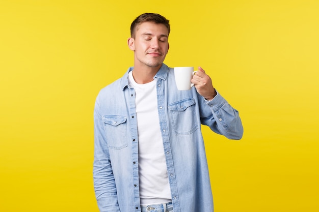 Estilo de vida, emociones de las personas y concepto de ocio. Hombre guapo despreocupado disfrutando de un agradable olor a café recién hecho, sosteniendo la taza, bebiendo té con los ojos cerrados y sonriendo de placer, fondo amarillo.