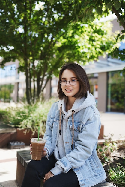 Estilo de vida fines de semana y concepto de personas Retrato de una joven caucásica con gafas de chaqueta de mezclilla mirando una cámara feliz sentada en un banco del parque con café bebiendo café con leche helada disfrutando de la primavera