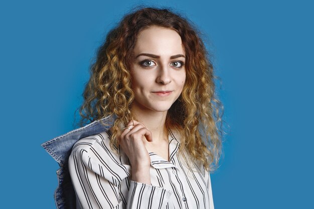 Estilo de vida y moda modernos. Mujer joven positiva con cara bonita y modelado de cabello voluminoso y ligero con elegante camisa a rayas y chaqueta de mezclilla en el hombro, marca de ropa publicitaria