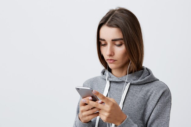 Estilo de vida moderno. Retrato de joven hermosa encantadora estudiante caucásica chica con cabello largo y oscuro con capucha gris mirando en teléfono inteligente con expresión seria y tranquila, mirando a través de las redes sociales