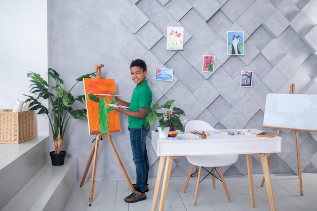 Estilo de vida. Perfil de un niño sonriente de piel oscura parado con paleta y pincel cerca del caballete mirando la cámara en una habitación moderna y luminosa con mesa y artículos de arte