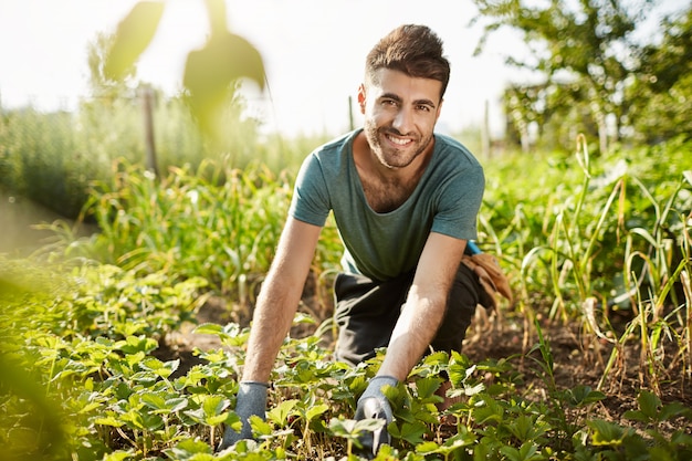 Estilo de vida saludable. La vida rural. Cerrar al aire libre retrato de joven agricultor caucásico barbudo atractivo sonriendo, pasando la mañana en el jardín cerca de la casa, recogiendo cultivos