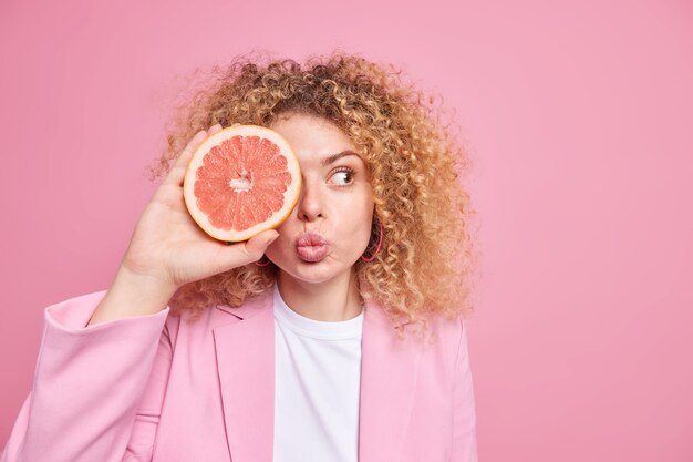 Estilo de vida saludable y vitaminas. Mujer encantadora tiene el pelo rizado y tupido mantiene los labios doblados cubre el ojo con la mitad de la toronja se mantiene a dieta vestida con ropa formal aislada sobre el espacio en blanco de la pared rosa