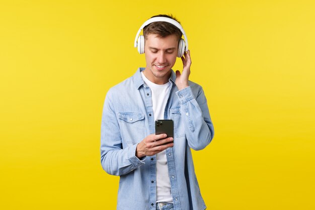 Estilo de vida, vacaciones de verano, concepto de tecnología. Apuesto joven moderno en ropa casual, escuchando música en auriculares, enviando mensajes con el teléfono inteligente, usando el teléfono móvil para hacer la lista de reproducción.