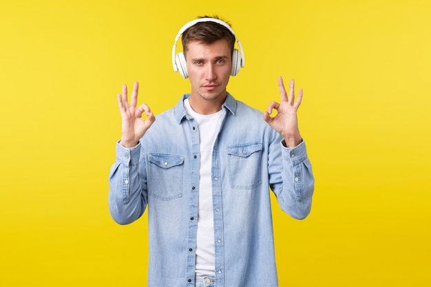 Estilo de vida, vacaciones de verano, concepto de tecnología. Cool guapo rubio en traje casual, mostrando un gesto bien, como una nueva canción impresionante o auriculares que compró en oferta, fondo amarillo.