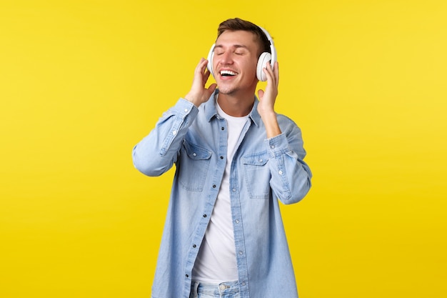 Estilo de vida, vacaciones de verano, concepto de tecnología. Hombre guapo feliz despreocupado cierra los ojos y se siente complacido, escucha la canción favorita en auriculares inalámbricos, siente la satisfacción del sonido perfecto.