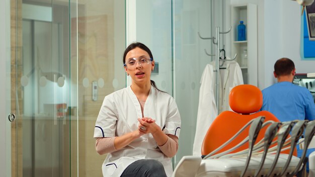 Estomatóloga mujer hablando por cámara de vídeo sentado en una silla en la clínica dental con asistente en segundo plano. Médico de odontología mirando por webcam explicando el tratamiento mientras la enfermera está hablando con el paciente