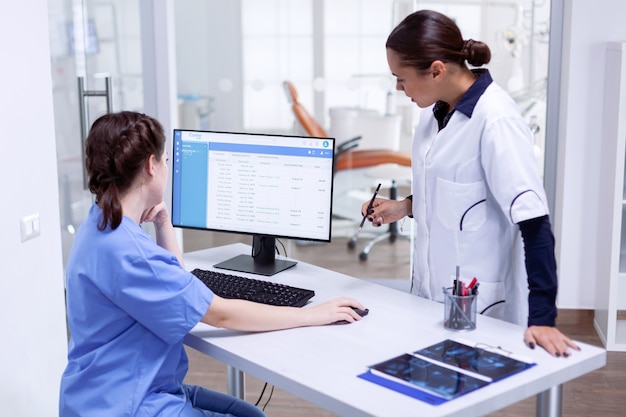 Estomatólogo y enfermera en la clínica dental que controla la cita del paciente mirando el monitor de la computadora. Asistente de estomatología y médico de dientes discutiendo en la recepción del consultorio dental