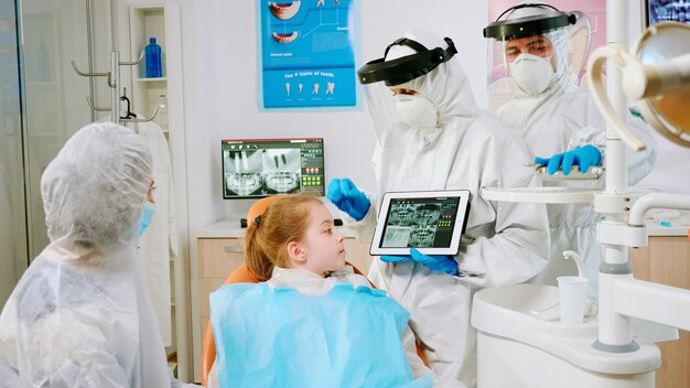 Estomatólogo en equipo de protección que muestra en la tableta de rayos x dentales revisándolo con la madre del paciente. Equipo médico con máscara de protección facial, guantes, explicando la radiografía usando la pantalla del portátil