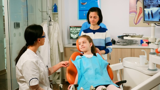 Estomatólogo explicando a la niña el proceso de limpieza de los dientes mientras el asistente del hombre prepara herramientas esterilizadas para su examen. Enfermera y médico trabajando juntos en la clínica estomatológica moderna