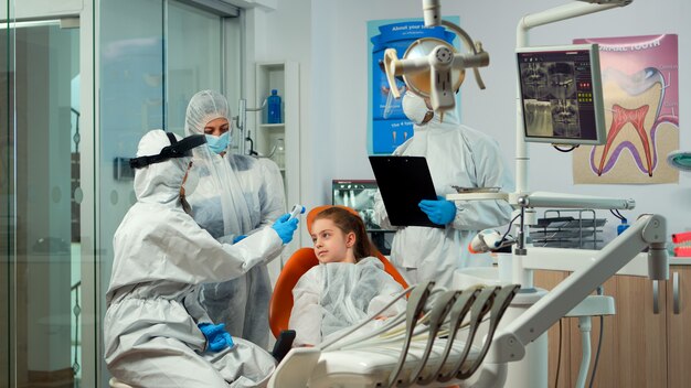 Estomatólogo con protector facial que mide la temperatura de la niña antes del examen dental durante la pandemia mundial. Concepto de nueva visita al dentista normal en el brote de coronavirus con traje protector.