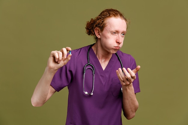 Estornudar sosteniendo pastillas joven médico masculino vistiendo uniforme con estetoscopio aislado sobre fondo verde