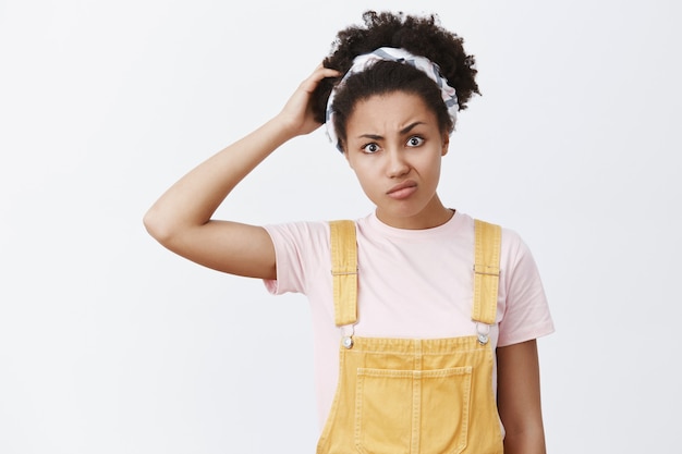 Estoy confundido. Retrato de mujer joven afroamericana linda dudosa insegura con overol amarillo y diadema, cabeza rasgada, frunciendo los labios y frunciendo el ceño mientras piensa, siendo interrogado e incierto