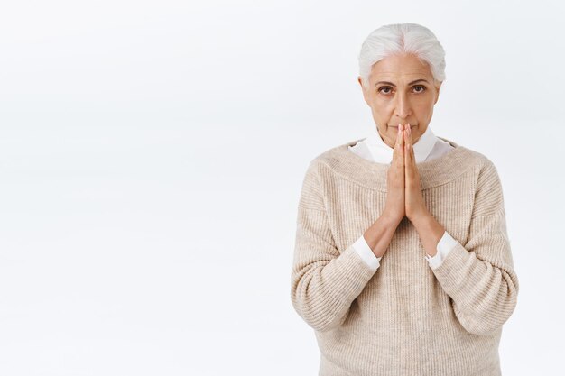 Estresada, preocupada elegante mujer mayor, anciana con cabello gris peinado, toca los labios con las manos en oración, buscando respuestas a Dios