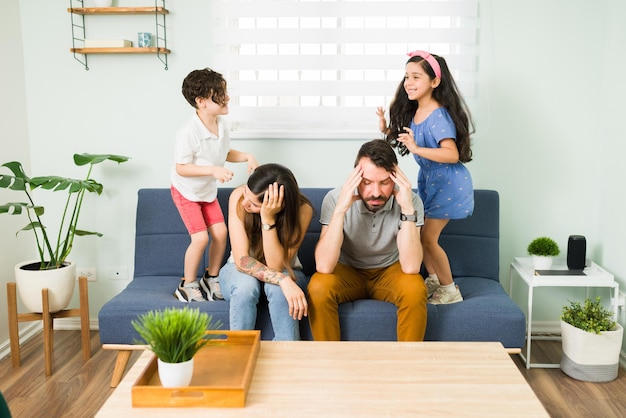 Estresados padres jóvenes molestos que se sienten cansados mientras sus hijos se portan mal y saltan en el sofá a su alrededor