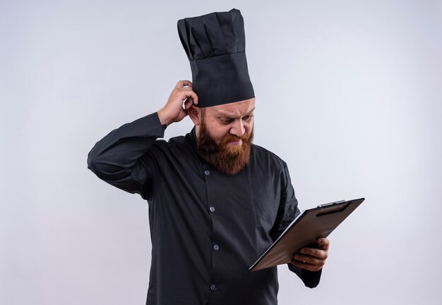 Un estresante chef barbudo con uniforme negro mirando una carpeta en blanco con la mano en la cabeza sobre una pared blanca