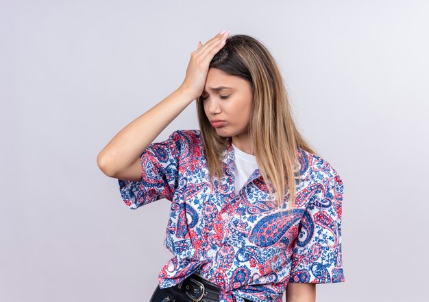 Una estresante joven y bella mujer vistiendo camisa estampada paisley manteniendo la mano en la cabeza sobre una pared blanca