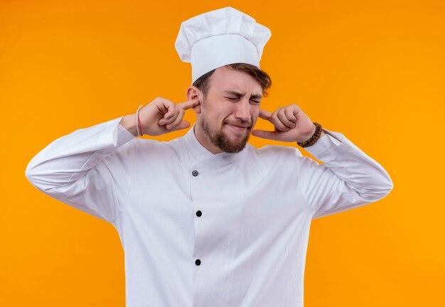 Un estresante joven chef barbudo en uniforme blanco sosteniendo las orejas con los dedos índices con los ojos cerrados en una pared naranja