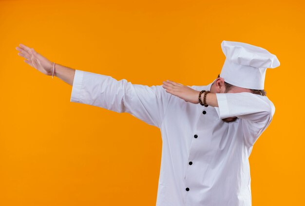 Un estresante joven chef barbudo en uniforme blanco sosteniendo sus manos sobre una pared naranja