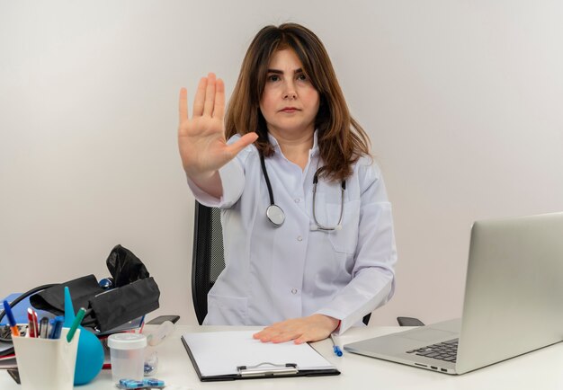 Estricta doctora de mediana edad con bata médica y estetoscopio sentado en el escritorio con portapapeles de herramientas médicas y portátil haciendo gesto de parada aislado