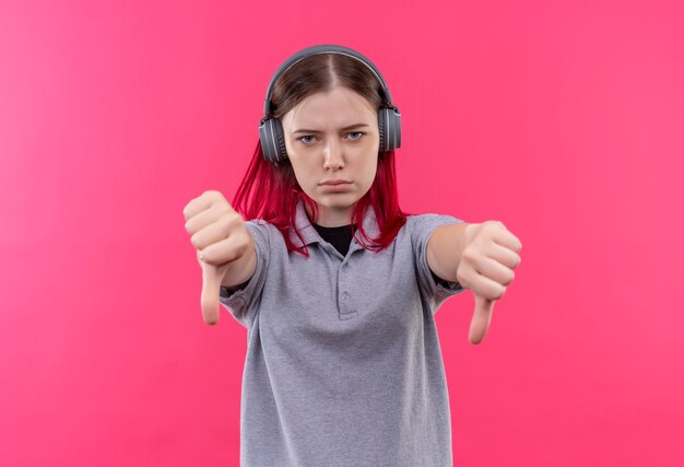 Estricta hermosa joven vestida con camiseta gris en auriculares con los pulgares hacia abajo sobre fondo rosa aislado