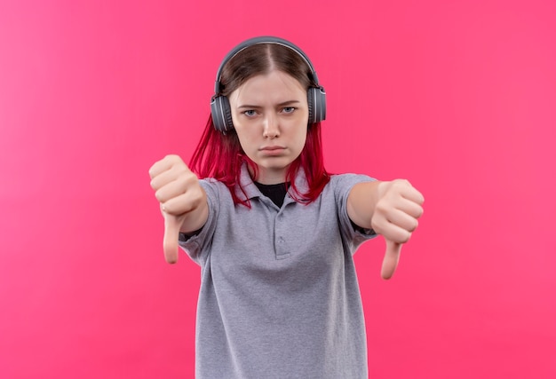 Estricta hermosa joven vestida con camiseta gris en auriculares con los pulgares hacia abajo sobre fondo rosa aislado