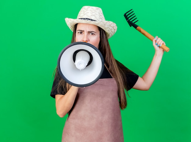 Estricta hermosa niña de jardinero en uniforme con sombrero de jardinería habla por altavoz y levantando rastrillo aislado en verde