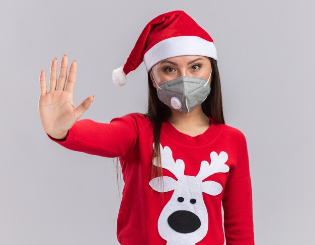 Estricta joven asiática con sombrero de navidad con suéter y máscara médica mostrando gesto de parada aislado sobre fondo blanco.