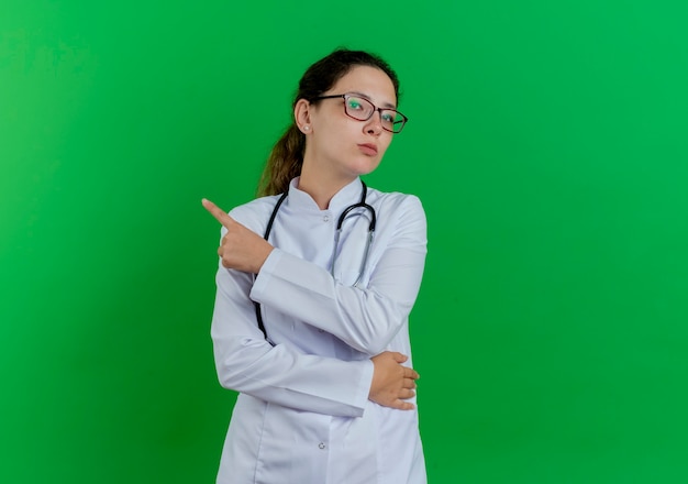 Estricta joven doctora vistiendo bata médica y estetoscopio y gafas apuntando al lado aislado en la pared verde con espacio de copia