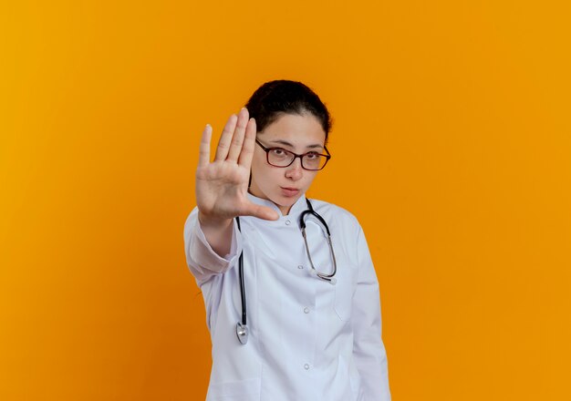 Estricta joven doctora vistiendo bata médica y estetoscopio con gafas mostrando gesto de parada aislado