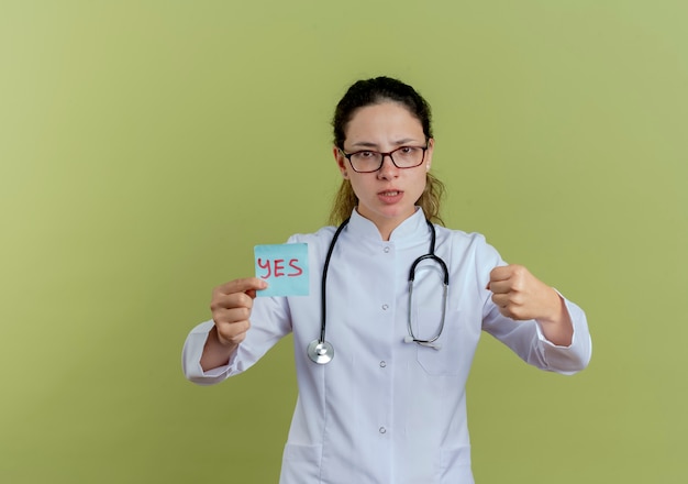 Estricta joven doctora vistiendo bata médica y estetoscopio con gafas sosteniendo una nota de papel que muestra el puño aislado