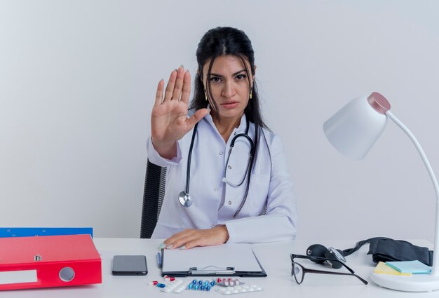 Estricta joven doctora vistiendo bata médica y un estetoscopio sentados en el escritorio con herramientas médicas poniendo la mano sobre el escritorio mirando haciendo gesto de parada aislado