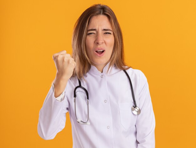 Estricta joven doctora vistiendo bata médica con estetoscopio sosteniendo el puño aislado en la pared amarilla