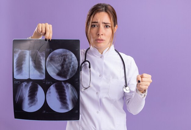 Estricta joven doctora vistiendo bata médica con estetoscopio sosteniendo rayos x y sosteniendo el puño aislado en la pared púrpura