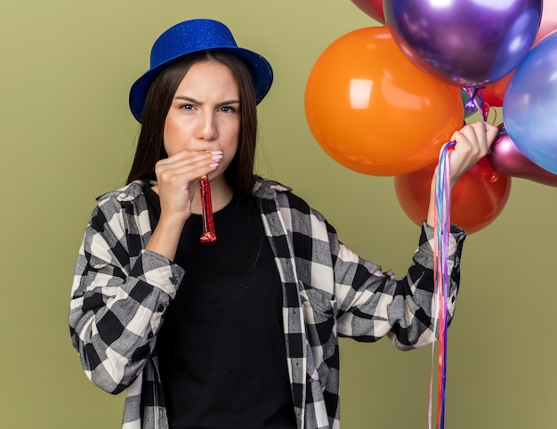 Estricta joven hermosa chica con sombrero azul sosteniendo globos que sopla el silbato del partido