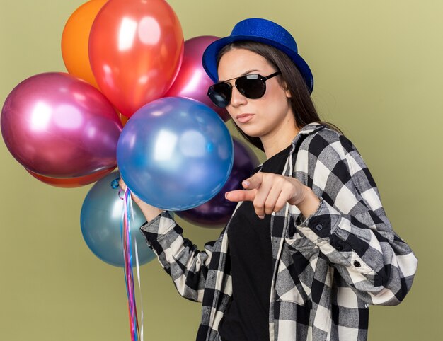 Foto gratuita estricta joven hermosa con sombrero azul con gafas sosteniendo globos puntos aislados en la pared verde oliva