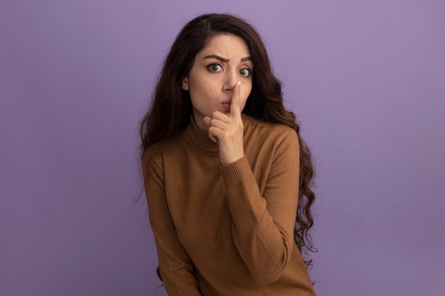 Estricta joven hermosa vestida con un suéter de cuello alto marrón que muestra el gesto de silencio aislado en la pared púrpura