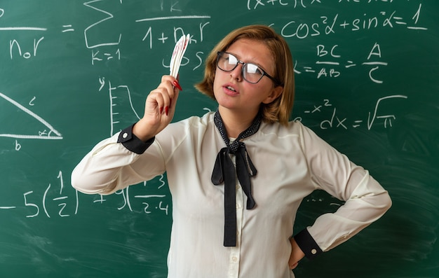 Estricta joven profesora con gafas de pie delante de la pizarra con número de ventiladores poniendo la mano en la cadera en el aula