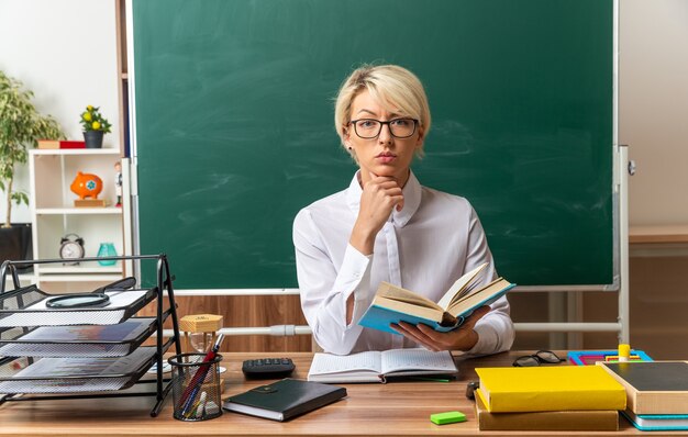 Estricta joven profesora rubia con gafas sentado en el escritorio con útiles escolares en el aula sosteniendo un libro abierto manteniendo la mano en la barbilla mirando al frente