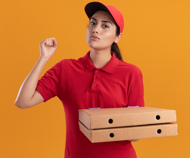 Estricta joven repartidora vestida con uniforme y gorra sosteniendo cajas de pizza levantando el puño aislado en la pared naranja