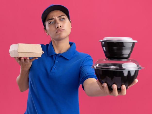 Estricta joven repartidora vestida con uniforme con gorra sosteniendo contenedores de comida aislados en la pared rosa