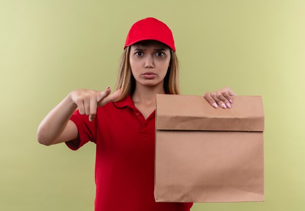 Foto gratuita estricta joven repartidora vestida con uniforme rojo y gorra sosteniendo una bolsa de papel y mostrando gesto aislado verde