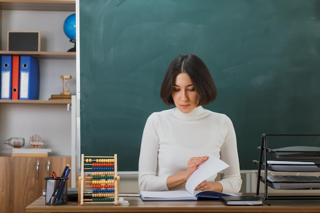 Estricta maestra joven volteando el libro sentada en el escritorio con herramientas escolares en el aula