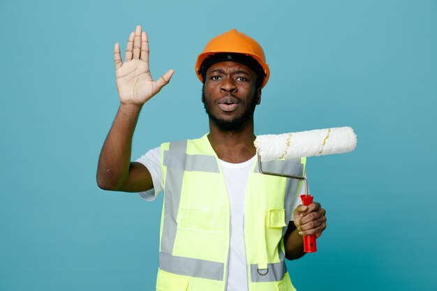 Estricta mano levantada joven constructor afroamericano en uniforme sosteniendo cepillo de rodillos aislado sobre fondo azul.