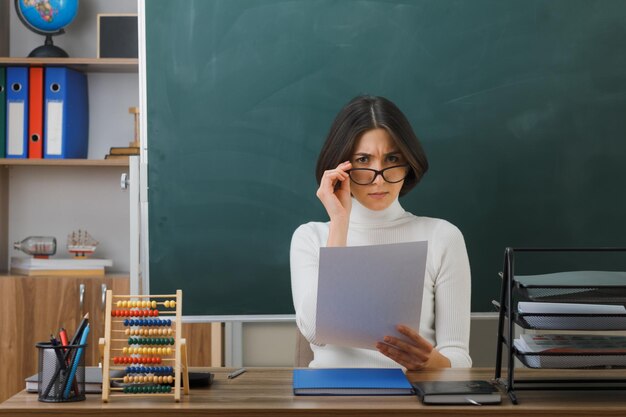 estricta mirando a la cámara joven maestra con gafas sosteniendo papel sentado en el escritorio con herramientas escolares en el aula